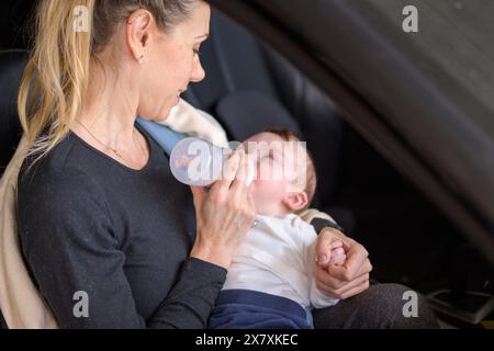 Donna bionda di mezza età che dà al suo bambino un biberon mentre è seduta in macchina in un garage Foto Stock