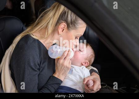 Donna bionda di mezza età che dà al suo bambino un biberon mentre è seduta in macchina in un garage Foto Stock