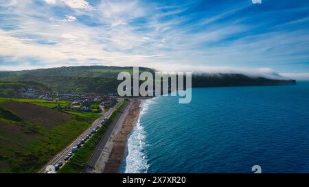 Vista aerea di un paesaggio costiero con una strada parallela al litorale, case, colline verdi e un orizzonte nebbioso sull'oceano. Foto Stock