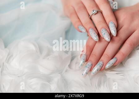 Vista ravvicinata di una persona mani elegantemente curate appoggiate su morbido tessuto bianco. Il design presenta un'intricata arte floreale blu accentuata da glit Foto Stock