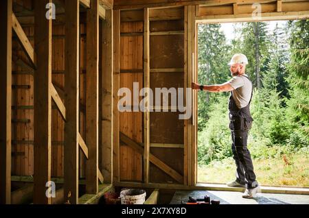 Falegname che costruisce una casa con struttura in legno adiacente alla foresta. L'uomo verifica la planarità delle pareti utilizzando una livella a bolla d'aria, indossata con indumenti protettivi, tra cui tuta e casco. Foto Stock