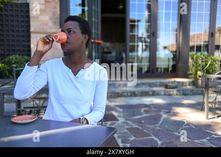 Una donna che sorseggia un caffè all'aperto su un patio, con finestre di vetro sullo sfondo, creando un'atmosfera tranquilla Foto Stock