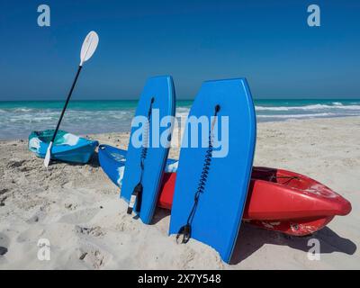 Due tavole blu, un kayak rosso e una pagaia su una spiaggia sabbiosa sotto un cielo blu, barche e pagaie su una spiaggia tropicale con acqua verde e blu sk Foto Stock