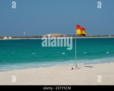 Bandiera rossa e gialla su una spiaggia di sabbia bianca con acque turchesi e cielo azzurro, mare verde scintillante con spiaggia, ombrelloni e lettini a fr Foto Stock