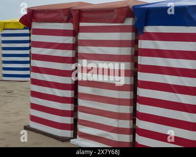 Capanne da spiaggia a righe in rosso-bianco e blu-bianco sulla spiaggia di sabbia, sedie a sdraio e tende da spiaggia in riva al mare sotto un cielo nuvoloso, passerella nella sabbia, Foto Stock