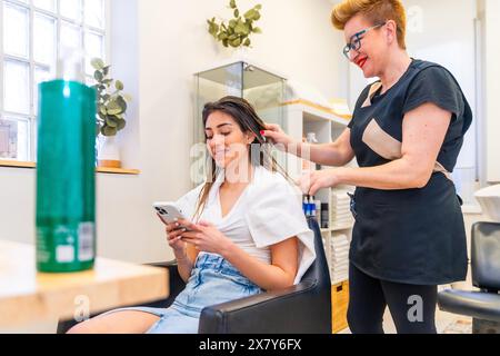 Parrucchiere pettinare i capelli di un cliente mentre utilizza il telefono cellulare Foto Stock