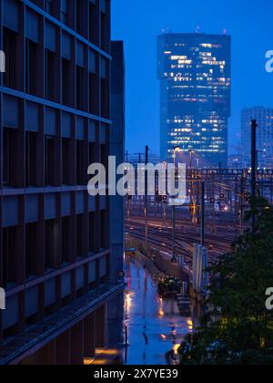 Zurigo, Svizzera - 16 maggio 2024: Scena urbana notturna con moderno edificio alto luminoso, strada bagnata e riflettente, luci per ufficio, cantiere ferroviario e blu a. Foto Stock