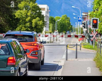 Schaan, Liechtenstein - 17 maggio 2024: Una scena suburbana con auto fermate ad un incrocio ferroviario, luci rosse accese, cancelli scendenti, con un SUV arancione e gree Foto Stock