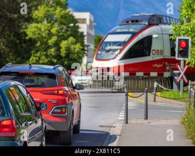 Schaan, Liechtenstein - 17 maggio 2024: Una scena suburbana con auto fermate ad un passaggio ferroviario, luci rosse accese, treno austriaco QBB, cancelli, con Foto Stock