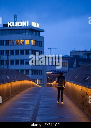 Zurigo, Svizzera - 16 maggio 2024: Persona con ombrello che cammina sul ponte illuminato al crepuscolo con l'edificio KUONI sullo sfondo. Foto Stock