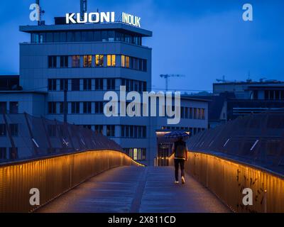 Zurigo, Svizzera - 16 maggio 2024: Persona con ombrello che cammina sul ponte illuminato al crepuscolo con l'edificio KUONI sullo sfondo. Foto Stock