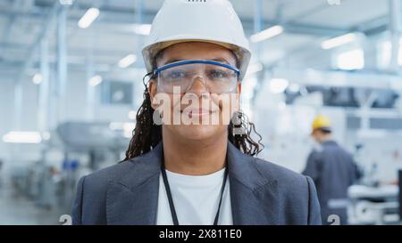 Primo piano ritratto di una donna africana di successo, felice e sorridente in bianco Hard Hat in piedi presso la fabbrica di produzione elettronica. Black Heavy Industry Specialist in posa per la fotocamera. Foto Stock