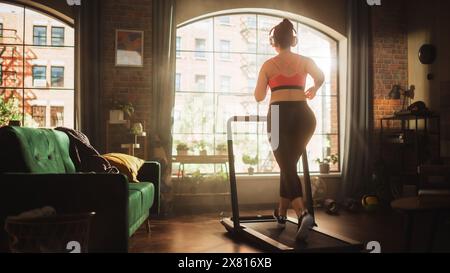 Bella atletica leggera Plus size Body positive Sports Woman che corre su un tapis roulant nella sua palestra di casa. Allenamenti di atleta femminile energica durante l'ascolto di podcast e musica in cuffia. Accogliente appartamento Foto Stock