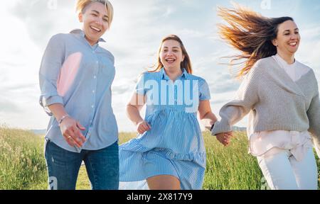 Ritratto di tre allegre donne sorridenti che corrono accanto al prato. Foto ad angolo basso sul cielo blu con nuvole. L'amicizia delle donne, le relazioni, e.. Foto Stock