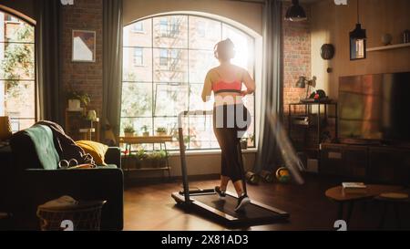 Bella atletica leggera Plus size Body positive Sports Woman che corre su un tapis roulant nella sua palestra di casa. Allenamenti di atleta femminile energica durante l'ascolto di podcast e musica in cuffia. Accogliente appartamento Foto Stock