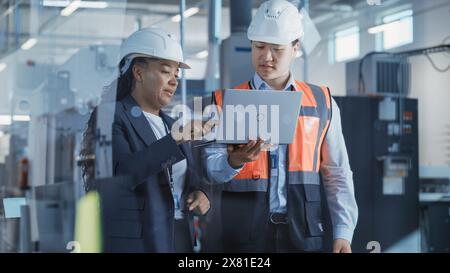 Due tecnici professionisti del settore pesante che indossano l'uniforme di sicurezza e i cappelli rigidi discutono del lavoro di fabbrica sui computer portatili. Specialista afroamericano e tecnico asiatico al lavoro. Foto Stock