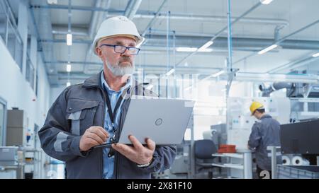 Ritratto di un direttore generale barbuto di mezza età in piedi in una fabbrica, con indosso una giacca da lavoro e un cappello bianco. Specialista del settore pesante che lavora su un notebook. Foto Stock