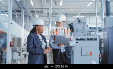 Due ingegneri professionisti del settore pesante che camminano in una fabbrica con uniforme di sicurezza e Hard Hats, discutendo del lavoro sui computer portatili. Specialista asiatico e tecnico afroamericano al lavoro. Foto Stock