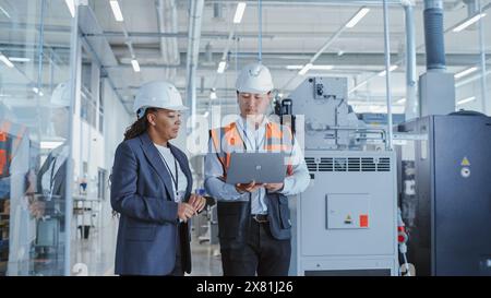 Due ingegneri professionisti del settore pesante che camminano in una fabbrica con uniforme di sicurezza e Hard Hats, discutendo del lavoro sui computer portatili. Specialista asiatico e tecnico afroamericano al lavoro. Foto Stock