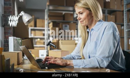 Felice responsabile dell'assistenza clienti che utilizza un computer portatile per controllare il numero d'ordine su un pacco, preparando una piccola scatola di cartone per la spedizione. Donna bionda proprietaria di una piccola impresa che lavora in magazzino. Foto Stock