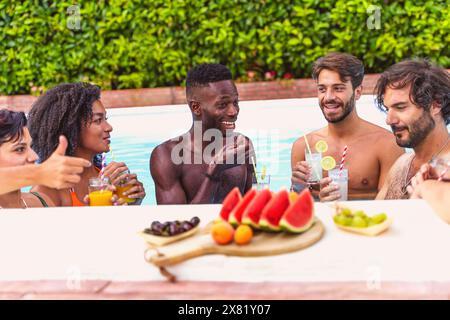 Amici multietnici che si godono un drink a bordo piscina - avendo un momento di relax e gioia - sorridendo e chiacchierando - incontro informale, felicità e amicizia Foto Stock
