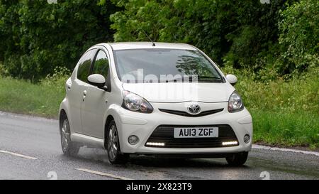 Stony Stratford, Regno Unito - 22 maggio 2024: 2013 Toyota Aygo bianca che guida su una strada britannica in una giornata umida Foto Stock