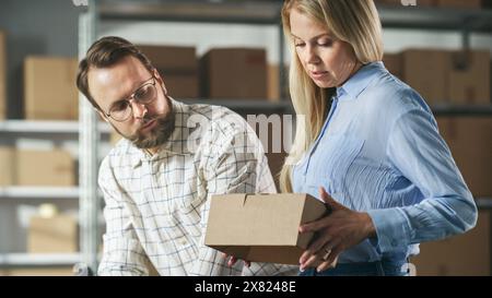 Responsabili dell'inventario del magazzino di uomini e donne che parlano, utilizzano un computer portatile e controllano le scorte al dettaglio. File di ripiani pieni di confezioni di cartone sullo sfondo. Foto Stock