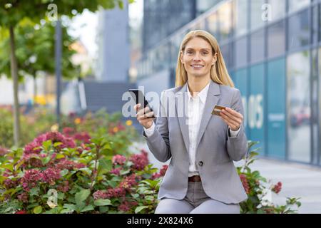 Una donna d'affari di successo, vestita con una tuta formale, sembra sicura di sé mentre tiene in mano una carta di credito e uno smartphone all'esterno di un moderno edificio per uffici circondato da vegetazione e fiori Foto Stock