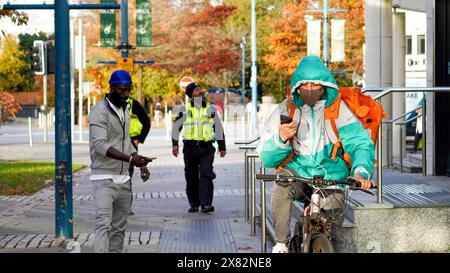 Cardiff, Galles, ottobre 29 2023: Uomo che guarda il suo telefono sulla sua bici che consegna cibo da asporto. Gli agenti di polizia stanno pattugliando le strade dietro. Foto Stock