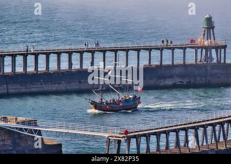 Un veliero in stile vintage che naviga attraverso un porto con un molo e un faro sullo sfondo. La gente cammina sul molo, e l'acqua io Foto Stock