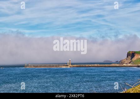 Una vista panoramica di un faro su un molo che si estende nell'oceano, con una scogliera sulla destra e un cielo nebbioso. Foto Stock