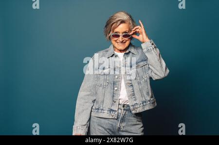 Elegante senior con capelli grigi e occhiali da vista si staglia giocosamente in uno studio indossando un outfit casual in denim. Foto Stock