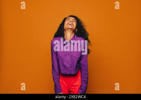 Donna allegra con capelli ricci su un vivace sfondo arancione. Indossando un abbigliamento casual elegante, irradia emozioni, felicità e divertimento. Foto Stock