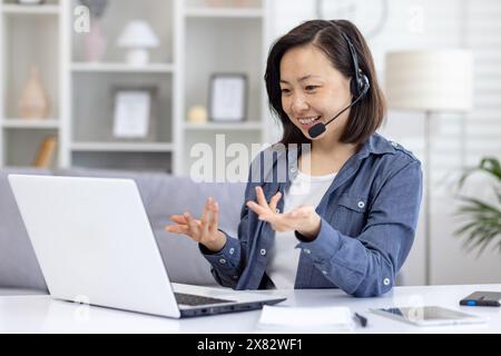Una donna sorridente che indossa un auricolare partecipa a una videochiamata sul proprio computer portatile mentre lavora da casa in un comodo salotto. Foto Stock