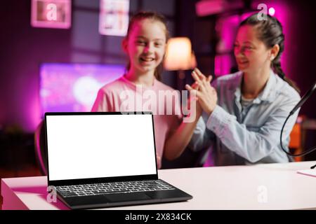 La ragazza allegra accanto a sua madre presenta un laptop in un salotto illuminato al neon rosa utilizzato come studio di vlogging. Giovane star dei media aiutato da un genitore a presentare un notebook con schermo isolato Foto Stock