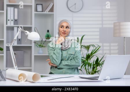 Giovane architetto donna che pensa all'interno dell'ufficio, donna in hijab che lavora con progetti e layout, utilizzando un notebook al lavoro. Foto Stock