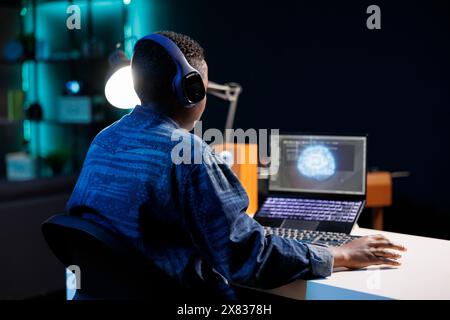 Il programmatore afroamericano con cuffie wireless sta lavorando sul suo computer portatile. Donna di colore che ascolta la musica mentre utilizza il codice e l'intelligenza artificiale per gestire e monitorare una rete neurale. Foto Stock