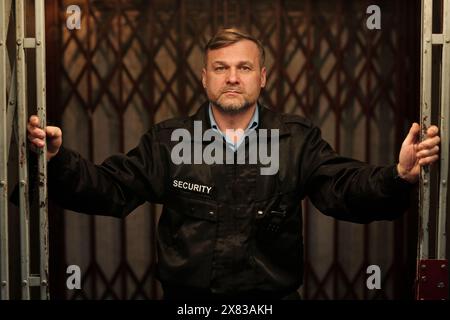 Guardia di sicurezza in uniforme nera che guarda la telecamera mentre apre o tiene il cancello a rete nel seminterrato del vecchio edificio al lavoro Foto Stock