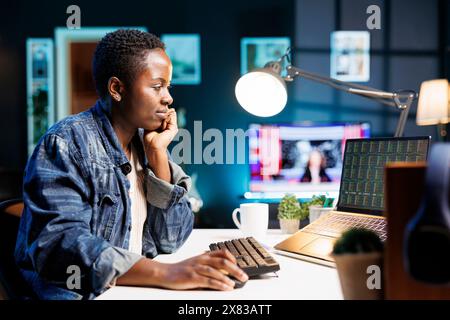 Il freelance afroamericano analizza attentamente i dati finanziari, confrontando le tendenze del mercato e prendendo decisioni commerciali strategiche online. Donna nera che si concentra sul notebook mostrando valori di tendenza commerciale. Foto Stock