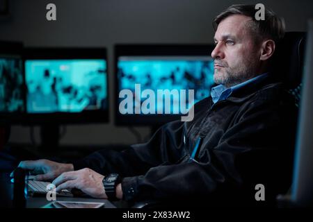 Guardia maschile matura in uniforme nera seduta accanto al posto di lavoro davanti al computer e guardando lo schermo con videocamera di videosorveglianza Foto Stock