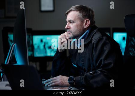 Serio guardia di sicurezza maschile che si concentra sulla visione di video CCTV sullo schermo del computer mentre è seduto accanto al posto di lavoro nella sala di osservazione Foto Stock