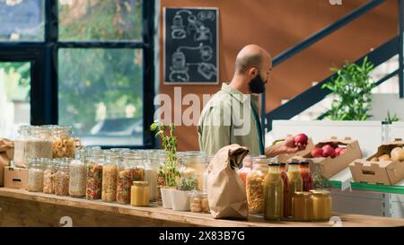 Il cliente entra nel negozio di alimentari locale per acquistare frutta e verdura biologica, guardando pasta e cereali conservati in vasetti di vetro sostenibili e non inquinanti. Uomo del Medio Oriente che pubblicizza una sana alimentazione. Foto Stock