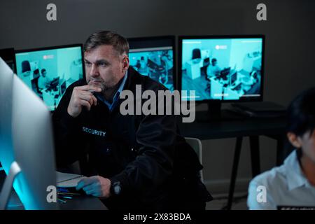 Guardia di sicurezza maschile matura che si concentra sulla visione di video cctv sullo schermo del computer mentre è seduto accanto al posto di lavoro nella sala di sorveglianza Foto Stock
