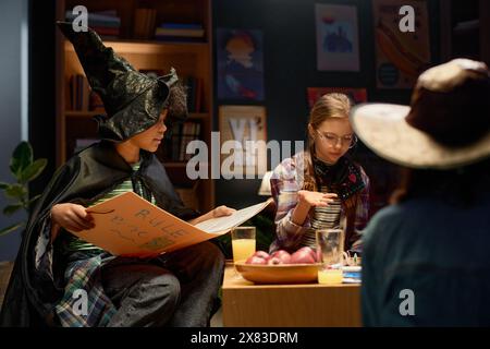 Simpatico ragazzo afroamericano con cappello da strega e mantello seduto accanto a una ragazza giovane durante una partita da tavolo e che spiega le regole del grande libro Foto Stock