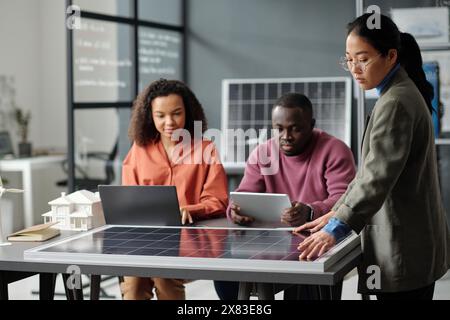 Giovane donna d'affari asiatica che tocca il modello del pannello solare mentre i suoi colleghi preparano nuovi progetti durante una riunione di lavoro Foto Stock