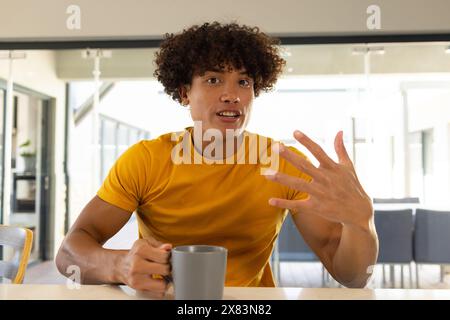 A casa, giovane birazziale in camicia gialla che chatta in videochiamata, tenendo in mano la tazza Foto Stock