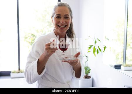 In bagno a casa, una donna caucasica matura che tiene una tazza di tè e sorride Foto Stock