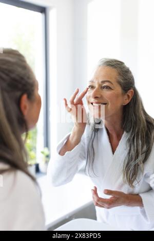 In bagno a casa, donna caucasica matura che applica crema sul viso Foto Stock