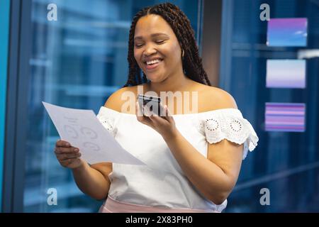 In un ufficio moderno per affari, giovane donna birazziale alla finestra con carta e smartphone Foto Stock