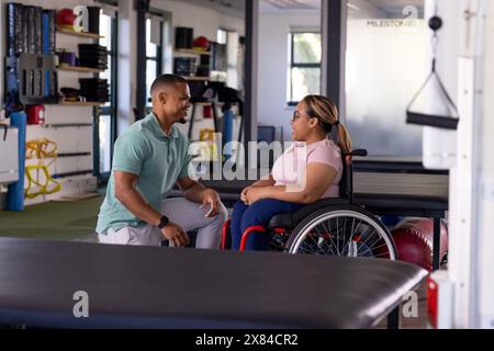 Un terapista maschile birazziale parla con una paziente paraplegica al centro di riabilitazione della palestra Foto Stock